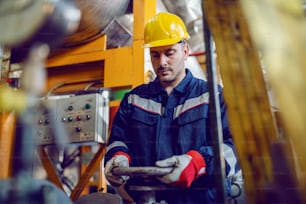 Hardworking energy plant worker in working suit and with protective helmet on head screwing valve.