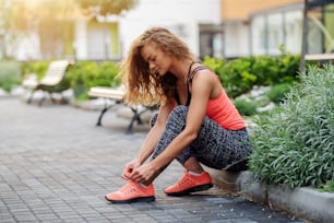 Charming fit caucasian woman dressed in sports wear sitting on roadside and tying shoelace. Park exterior.