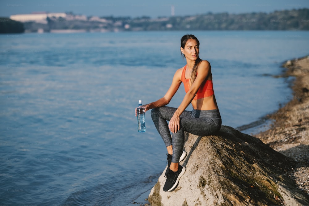 Fit slim attractive caucasian brunette in sportswear and with ponytail sitting on the rock near river, holding bottle with water, resting and looking away.