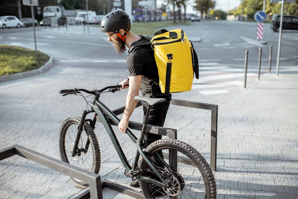 Jovem mensageiro barbudo entregando comida com saco térmico, estacionando sua bicicleta na rua perto do escritório. Conceito de serviço de entrega