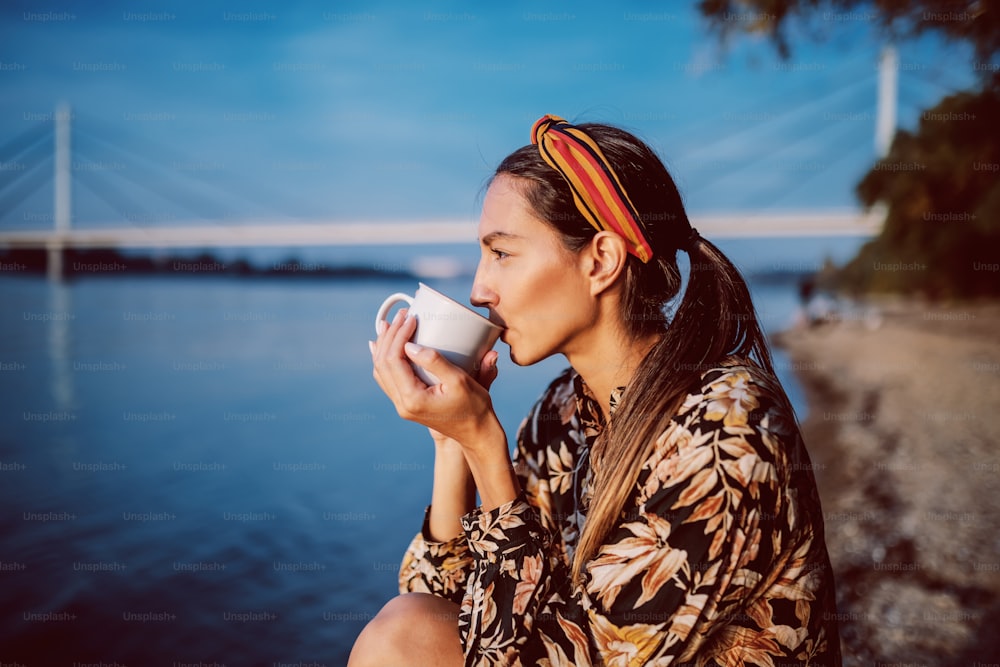 Side view of charming brunette in floral dress and with headband sitting on shore and drinking beverage from mug. In background is bridge.
