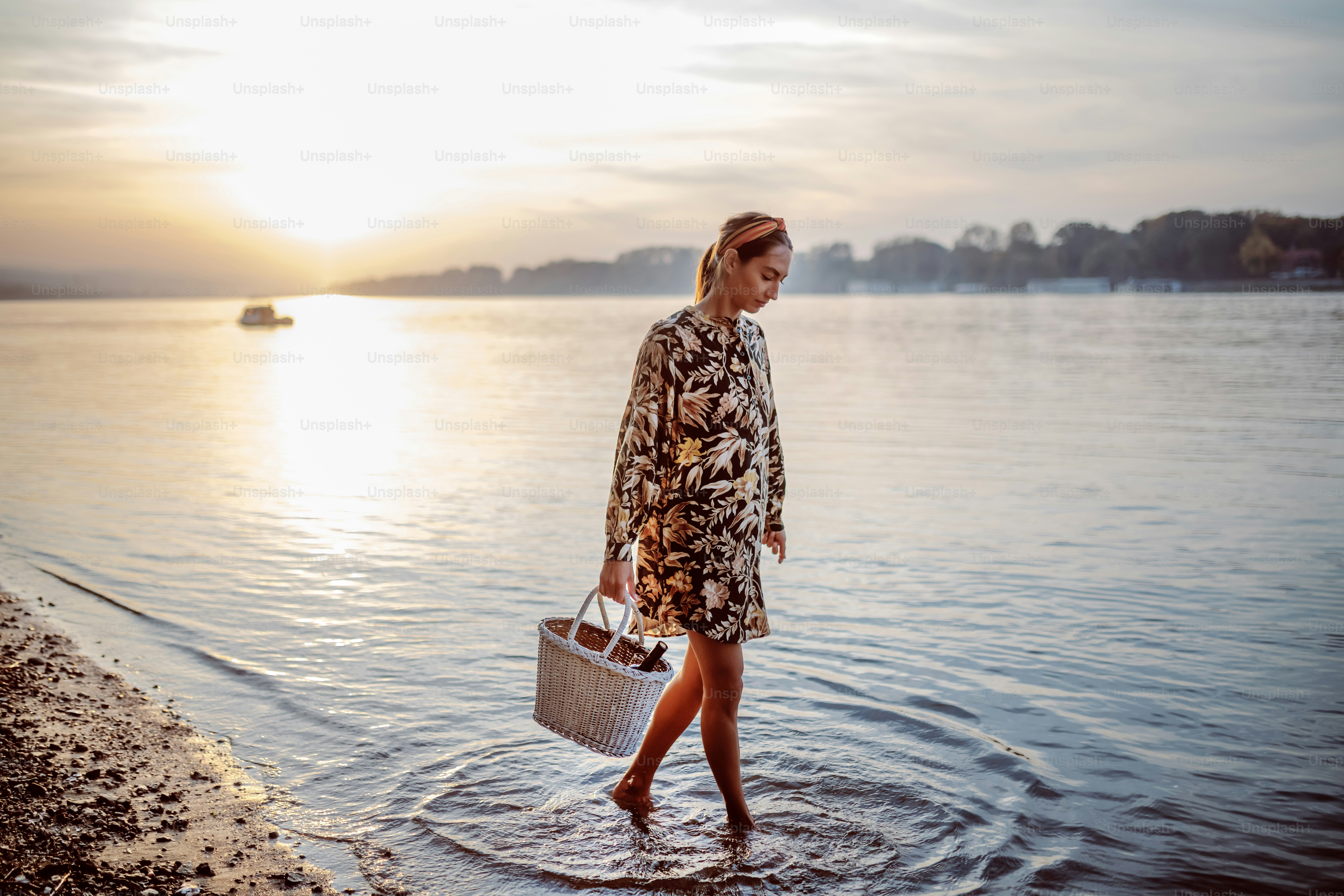 Full length of attractive caucasian brunette in floral dress and with headband walking in water and holding picnic basket. Summertime.