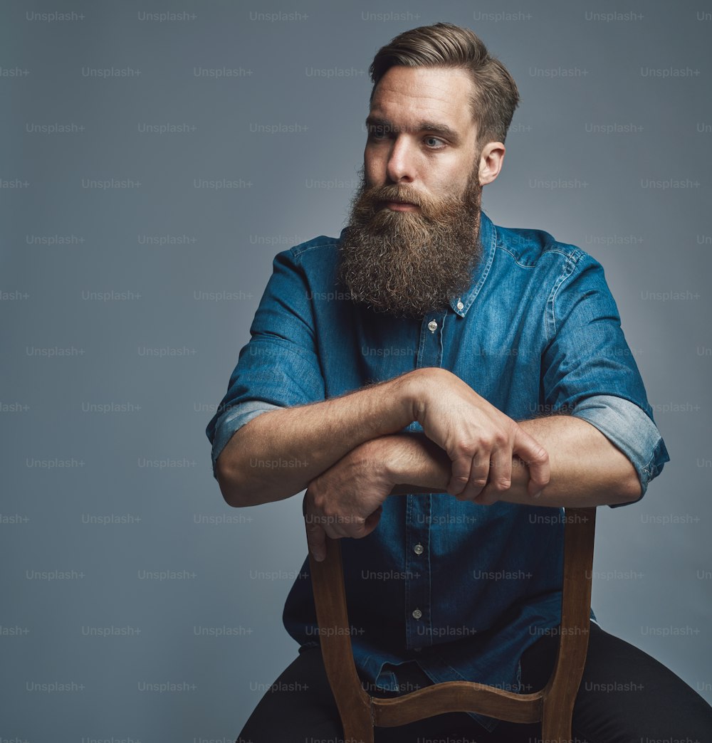 Hip and stylish young man with a long beard looking deep in thought while sitting in a chair against a gray background