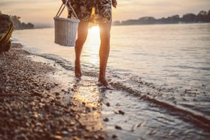 水中を歩く女性の足のトリミングされた写真�。ピクニックバスケットを手に持つ女性。背景には太陽が写っています。