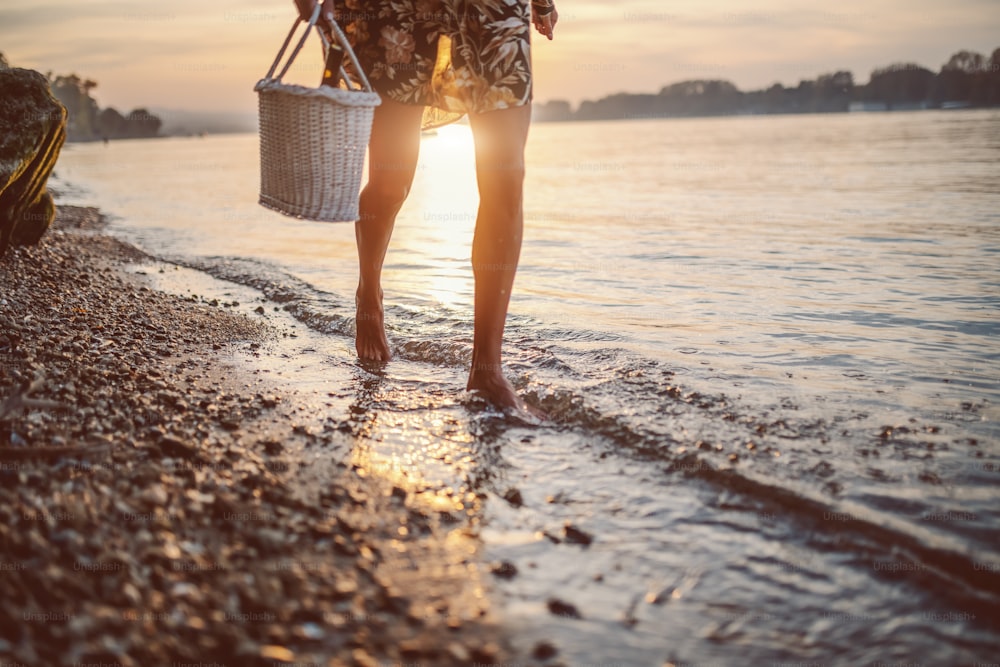 Cropped picture of female's legs walking in water. Woman holding picnic basket in hands. In background is sun.