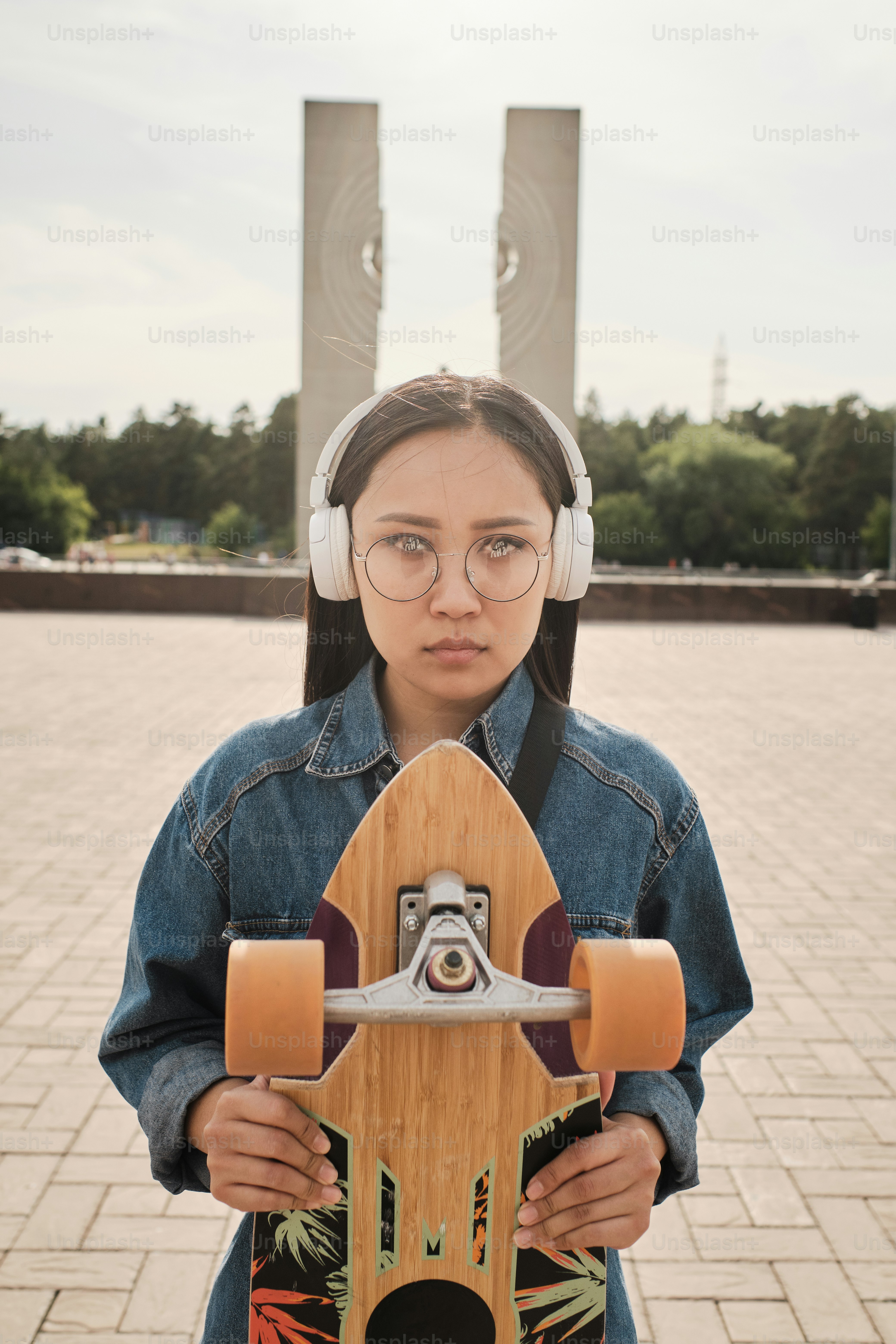Urban portrait of young asian skater girl standing outdoors, posing with her longboard, wearing white headphones, blue denim