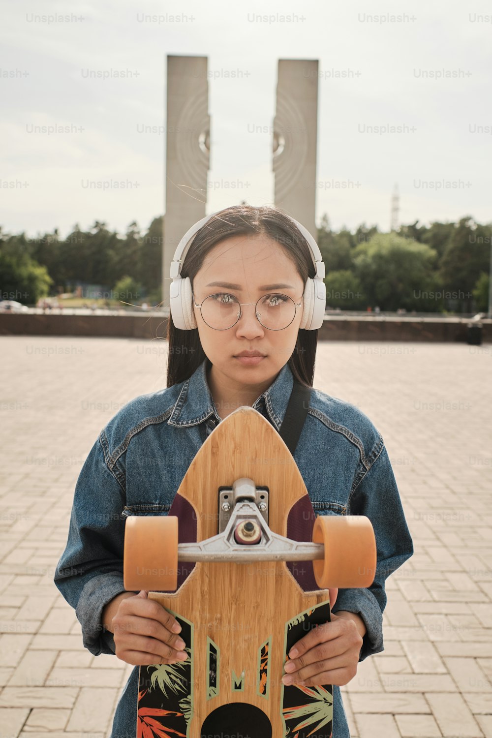 Urban portrait of young asian skater girl standing outdoors, posing with her longboard, wearing white headphones, blue denim