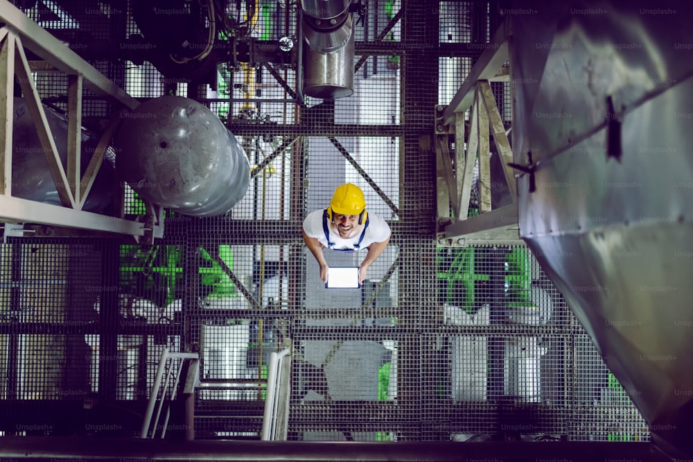 Vista superior do trabalhador da planta sorridente em roupas de proteção usando tablet.