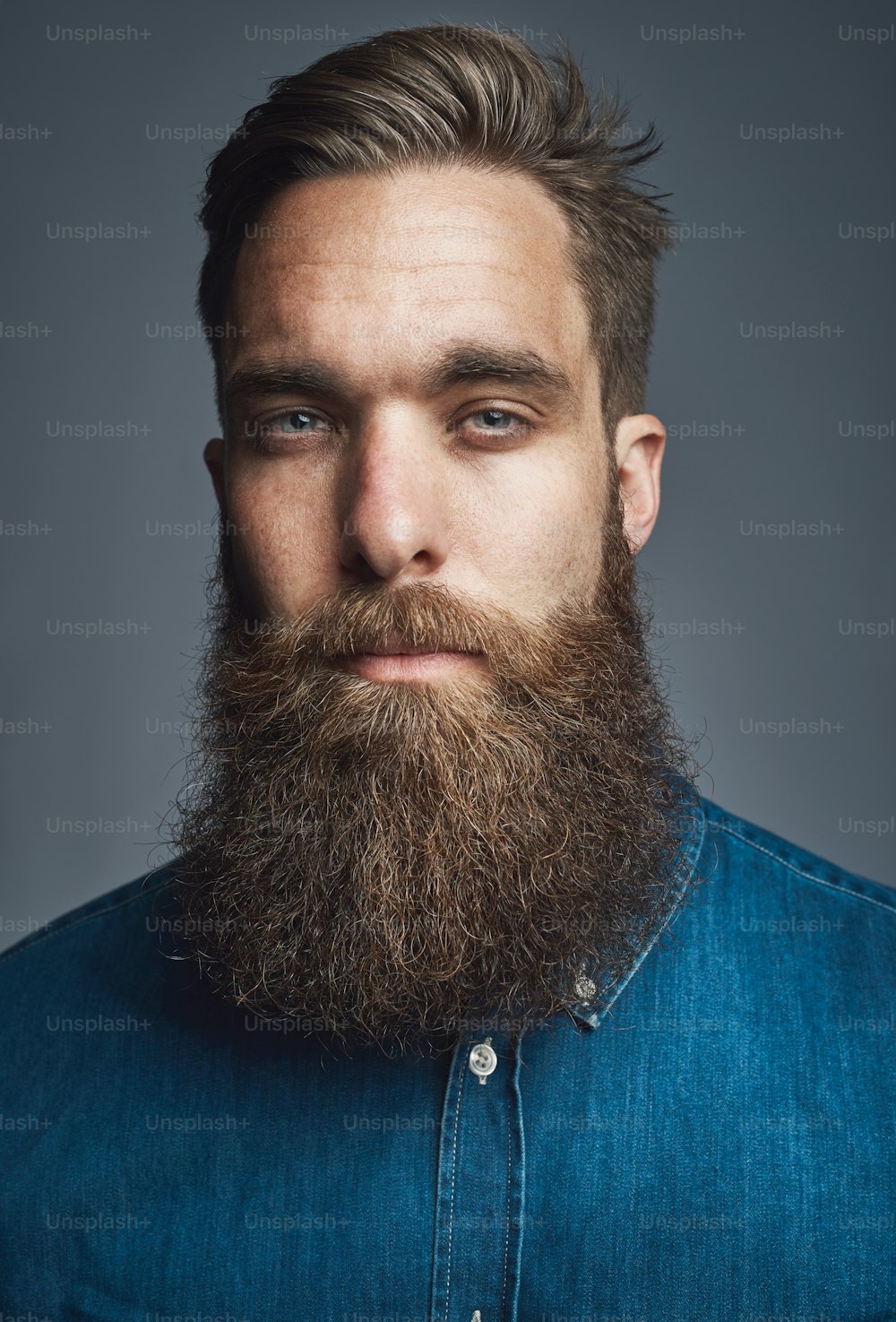 Gros plan d’un jeune homme élégant avec une longue barbe debout seul dans un studio sur un fond gris