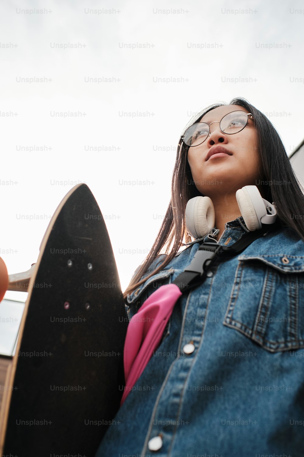 Portrait from below of young asian skater girl with her longboard, wearing round glasses, blue jeans and pink belt bag