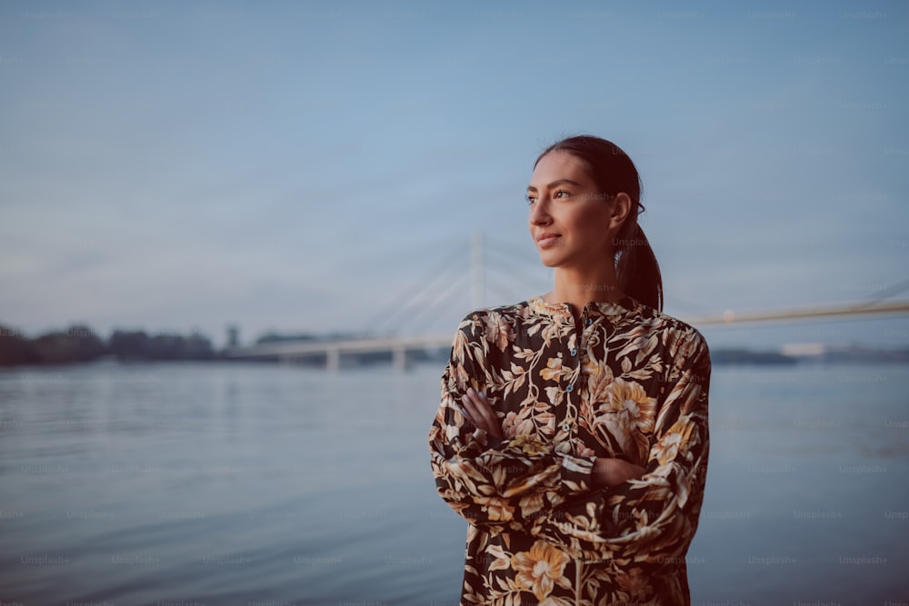 Beautiful caucasian brunette in floral dress standing on shore with hands folded and looking away. In background is bridge.