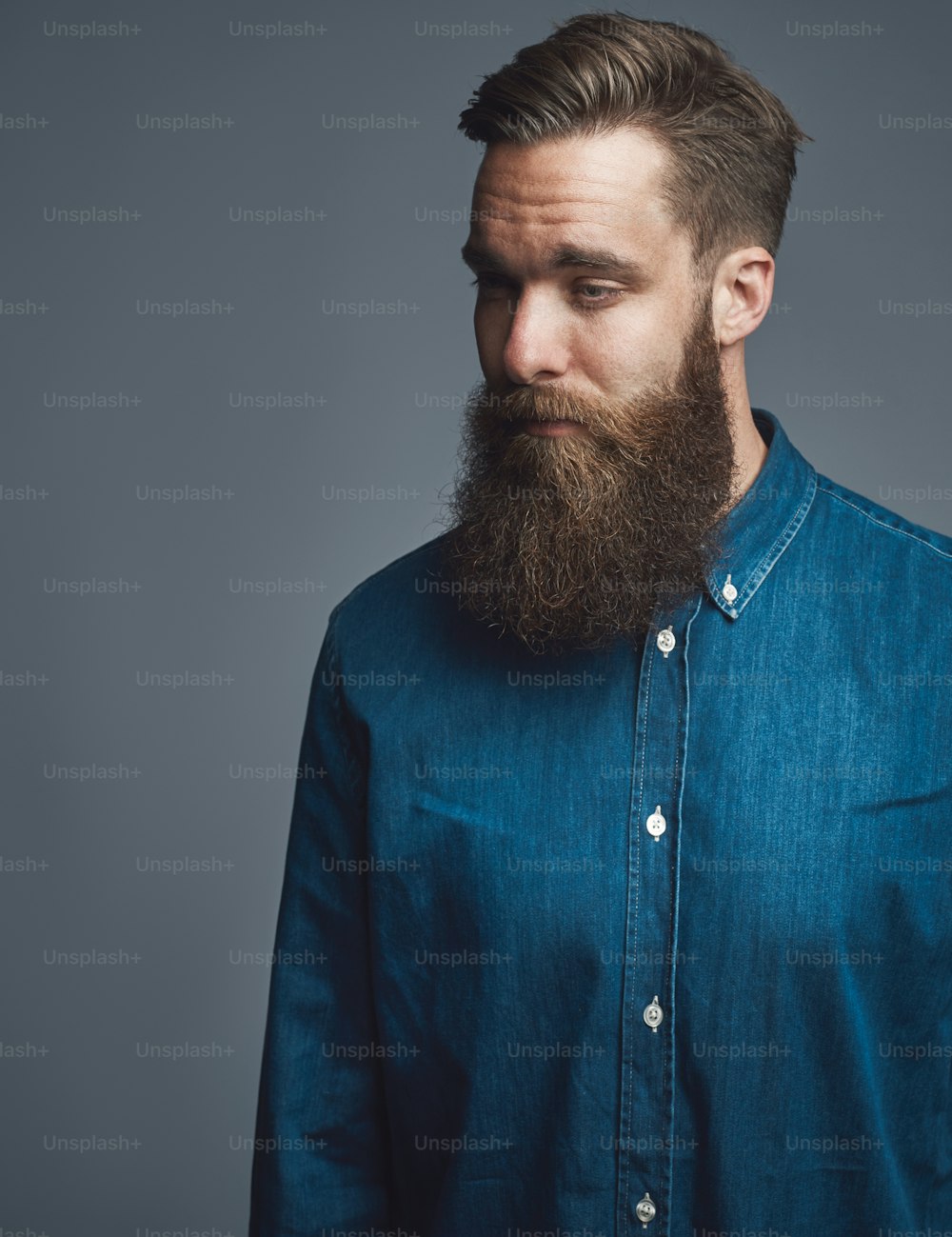 Stylish young man with a long beard looking deep in thought while standing alone against a gray background