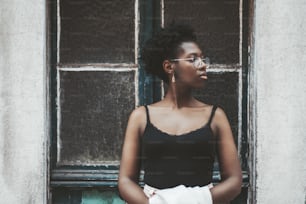 A charming young African female in spectacles is standing in front of an old antique wall with a doorway and  pensively looking aside; a charming black girl in eyeglasses outdoors near the door