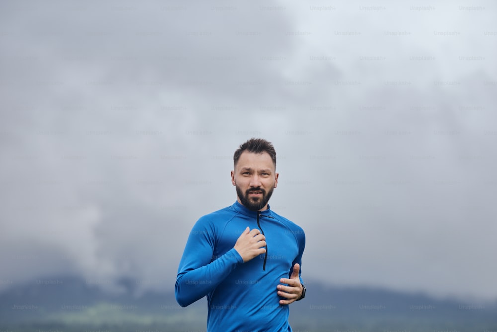 Mid age sportsman running outdoor in summer, on gloomy day, with scenic view, wearing blue shirt, he has a beard