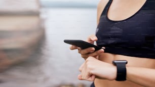 Close up of mid age skinny sports woman checking smart watch with smart phone, synchronizing, exercising outdoor in summer, on gloomy day with scenic view, wearing tank