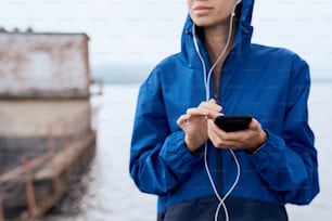 Close up of female hands holding smart phone with earphones outdoor in summer, on gloomy day with scenic view, wearing blue rain coat