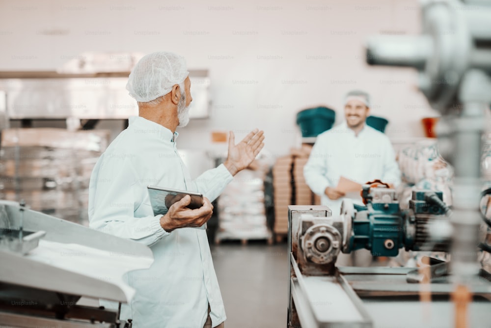 Senior adult Caucasian supervisor talking to his younger colleague about quality of food. Man holding tablet. Both are dressed in white uniforms and having hairnets. Food plant interior.