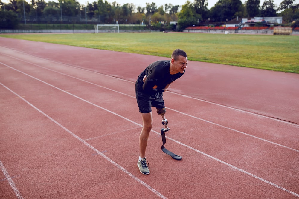 Sporty caucasian man with artificial leg standing on running track and having pain in stomach.