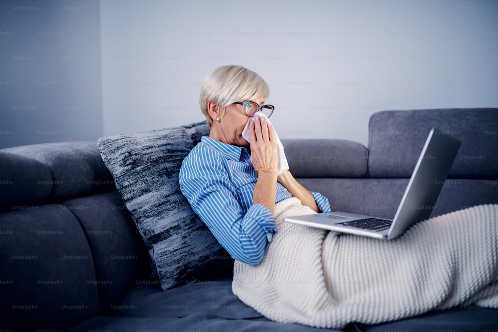 Sick caucasian senior woman sitting on sofa li living room covered with blanket and blowing nose while holding laptop in lap.