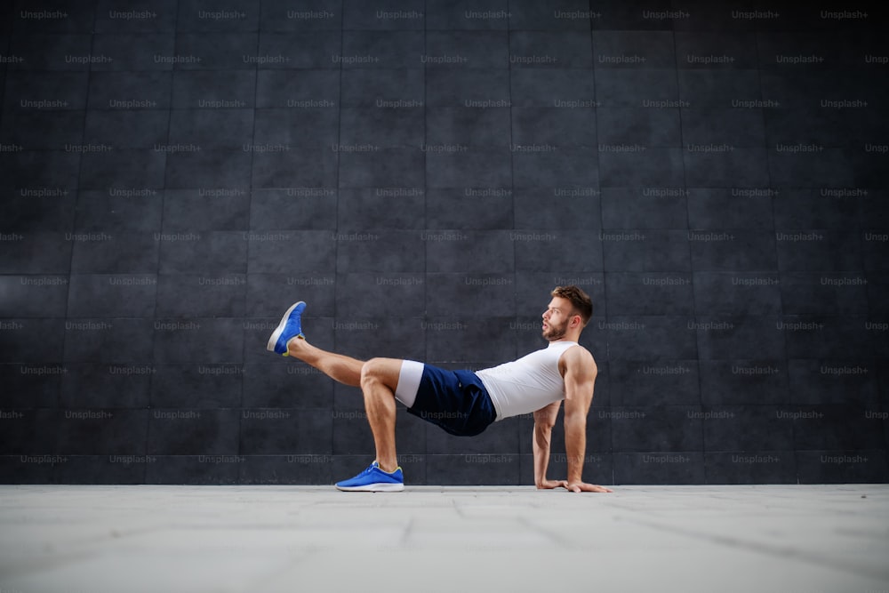 Handsome sporty Caucasian man in shorts doing forward kicks in front of gray background.