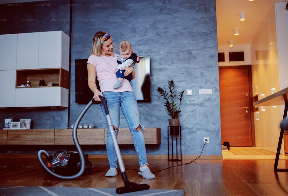 Worthy caucasian blond housewife holding her loving baby and using vacuum cleaner to clean floor in living room.