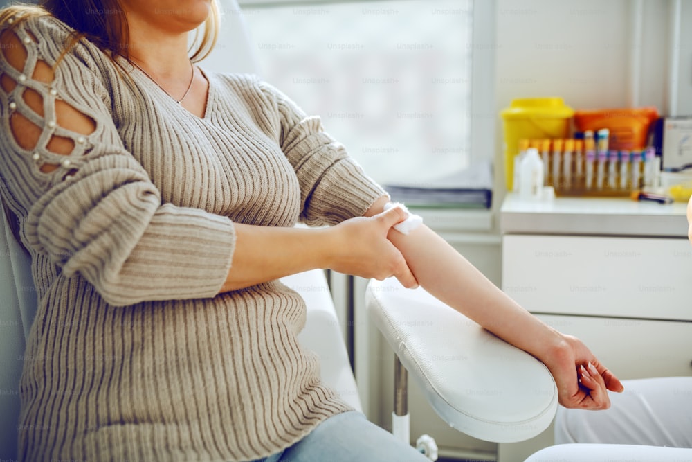Cropped picture of female patient sitting in chair in lab and holding absorbent cotton after taking blood sample from vein.