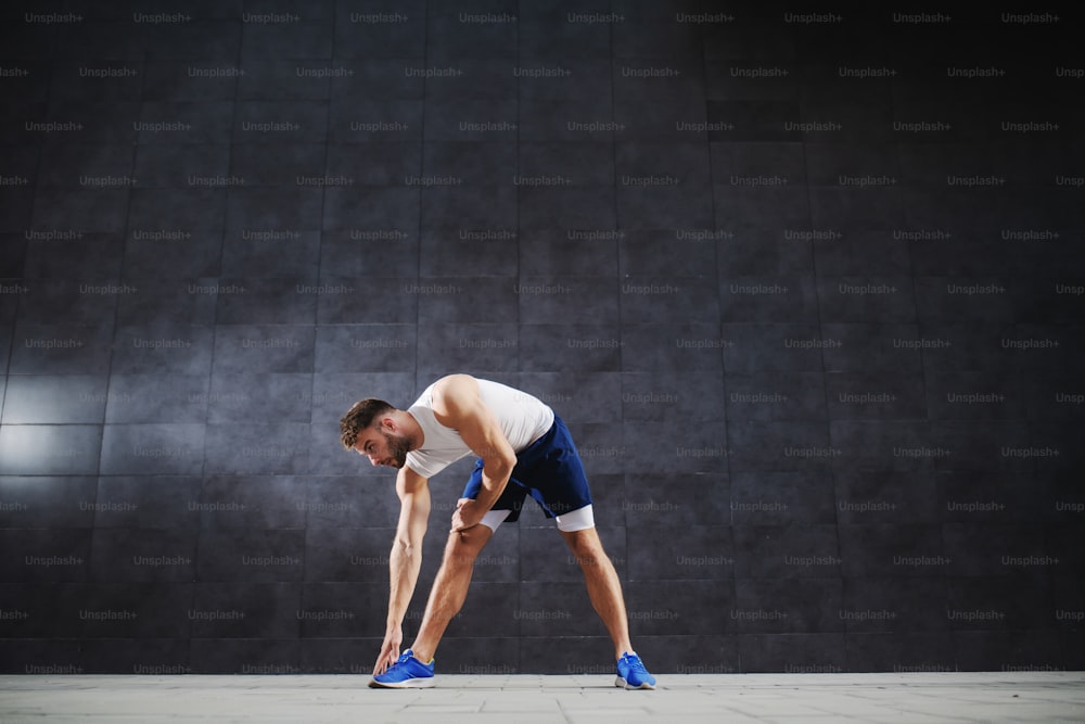 Sporty dedicated caucasian in shorts and t-shirt doing stretching exercises while standing outdoors in front of gray wall.