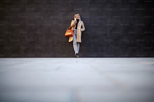 Full length of attractive caucasian smiling blonde woman in coat and handbag leaning on wall and talking on the smart phone.