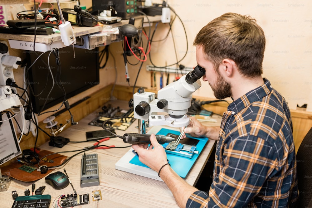 Junger bärtiger Reparateur mit elektrischem Handwerkzeug schaut während der Arbeit mit winzigen Details von kaputtem Gerät in der Werkstatt ins Mikroskop