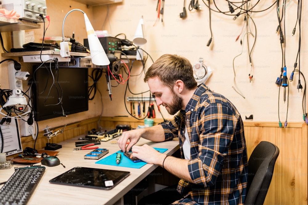 Professioneller Reparateur, der sich über demontiertes Smartphone beugt und winzige Details mit Hilfe einer Pinzette in der Werkstatt repariert