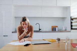 Cute caucasian blonde woman in sweater leaning on dining table and calculating bills. In hand is pen. Apartment interior.
