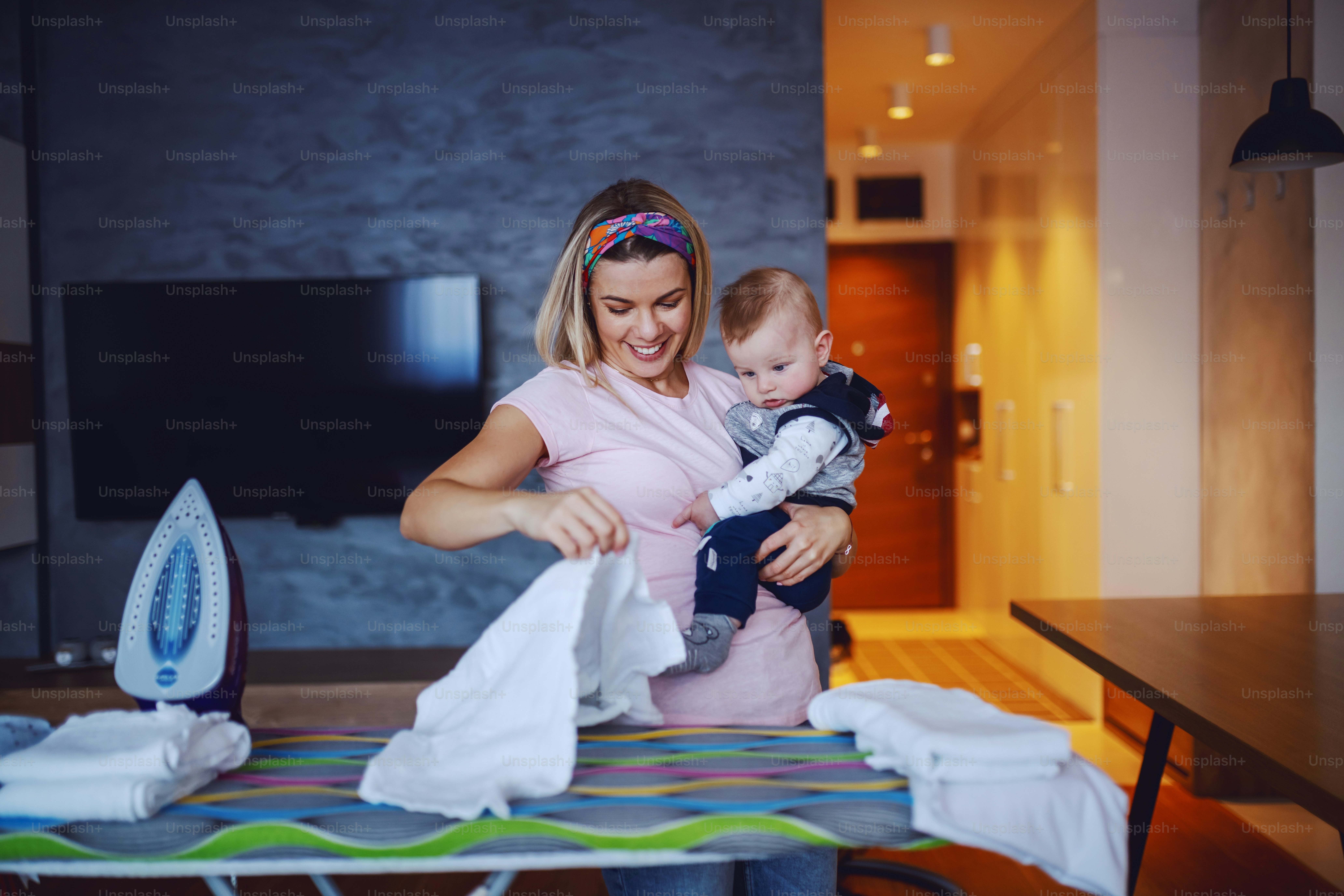 美しい白人のブロンドの主婦は、リビングルームに立って服にアイロンをかけ、赤ちゃんを抱いています。