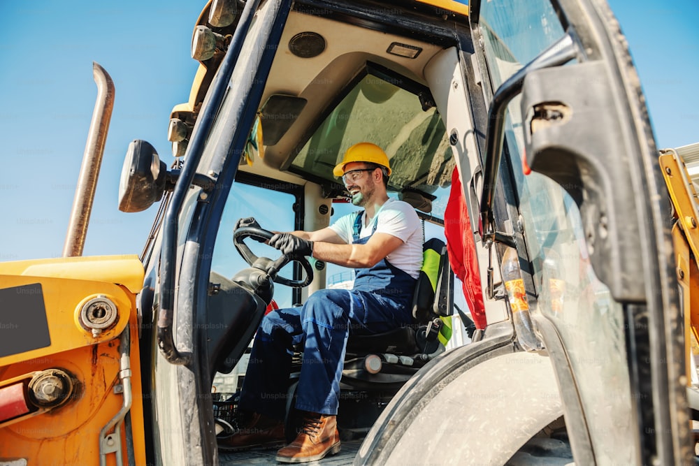 Smiling handsome caucasian worker in overall and with helmet on head driving excavator.