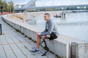 Guapo hombre discapacitado caucásico deportivo en ropa deportiva y con pierna artificial sentado en el muelle.