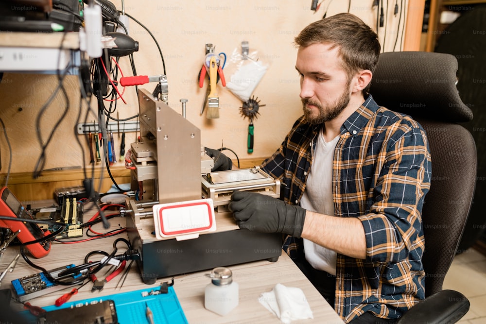 Bearded repairman in protective gloves sitting by working equipment while trying to find out trouble of broken smartphone