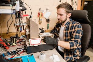 Bearded repairman in protective gloves sitting by working equipment while trying to find out trouble of broken smartphone