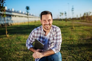 Handsome smiling caucasian farmer in plaid shirt and jeans crouching outdoors with tablet in hands. In background are barn and trees.
