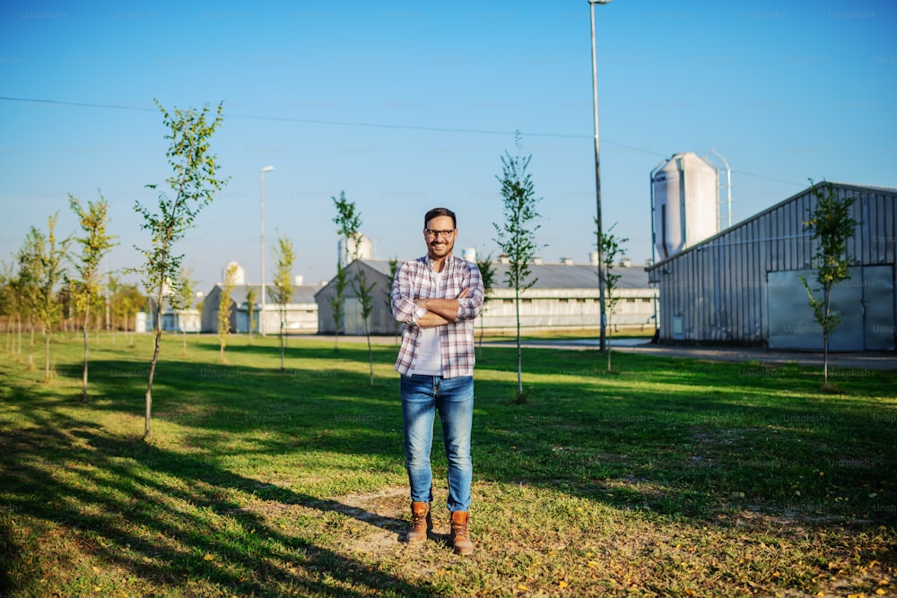 Guapo granjero caucásico sonriente con camisa a cuadros y jeans parado al aire libre con los brazos cruzados. Al fondo están los graneros.