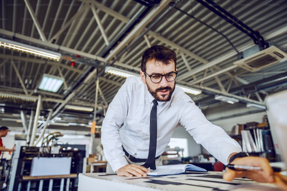 Handsome caucasian bearded supervisor with eyeglasses and in shirt and tie checking on quality of printed sheets while standing in printing shop.