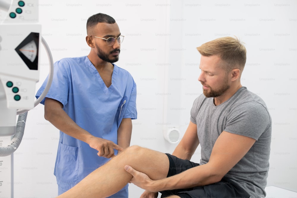 Young doctor in uniform consulting sportsman while pointing at his sick leg or knee during visit in hospital