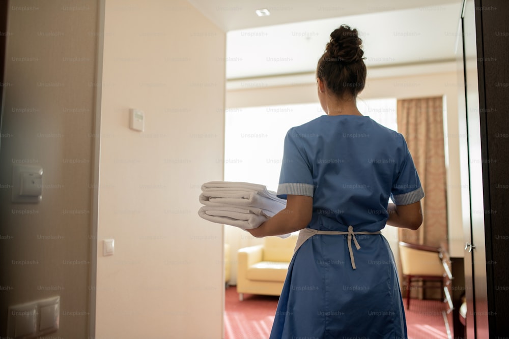 Vista trasera de la joven camarera morena en uniforme que lleva una pila de toallas blancas limpias para los invitados al entrar en una de las habitaciones