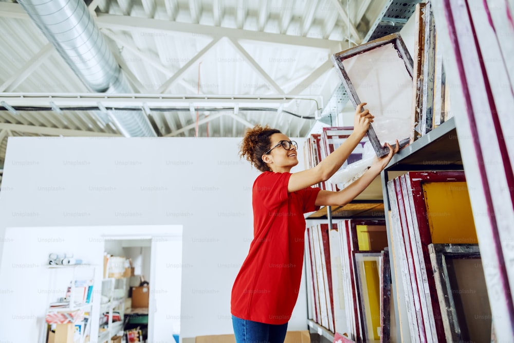 Cute caucasian female employee with curly hair and with eyeglasses reaching for silk screen plates. Printing shop interior.