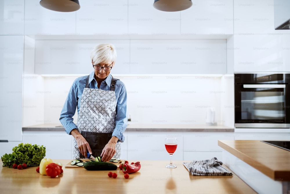 Donna anziana caucasica sorridente in grembiule in piedi in cucina e taglio cetriolo. Sul bancone della cucina ci sono peperoni, lattuga e un bicchiere di vino.