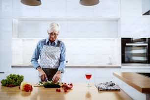 Mulher idosa caucasiana sorridente no avental em pé na cozinha e cortando pepino. No balcão da cozinha estão pimentos, alface e copo de vinho.