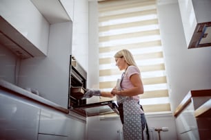 Vista angolare bassa dell'attraente casalinga bionda caucasica in grembiule che tira fuori la cena al forno dal forno.