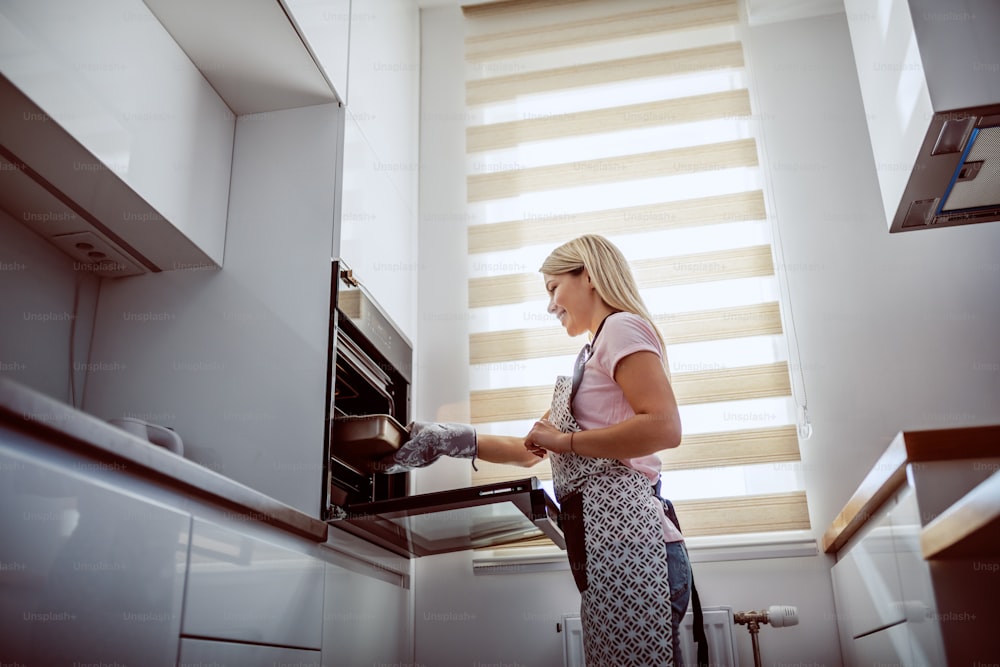 Low angle view of attractive caucasian blond housewife in apron taking out baked dinner from oven.