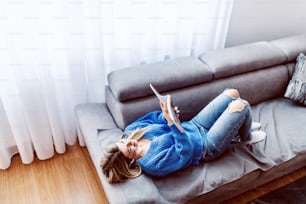 Top view of attractive caucasian blonde woman lying on sofa in living room, relaxing and reading interesting book.