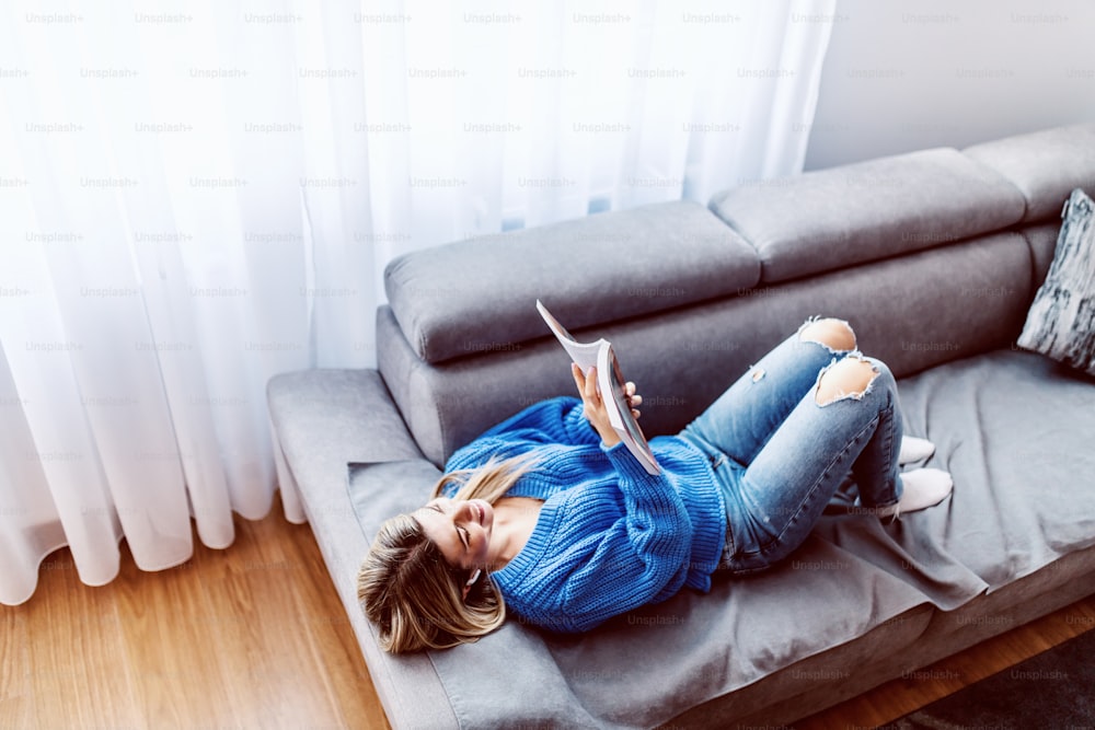 Vista dall'alto di attraente donna bionda caucasica sdraiata sul divano in soggiorno, rilassante e leggendo un libro interessante.
