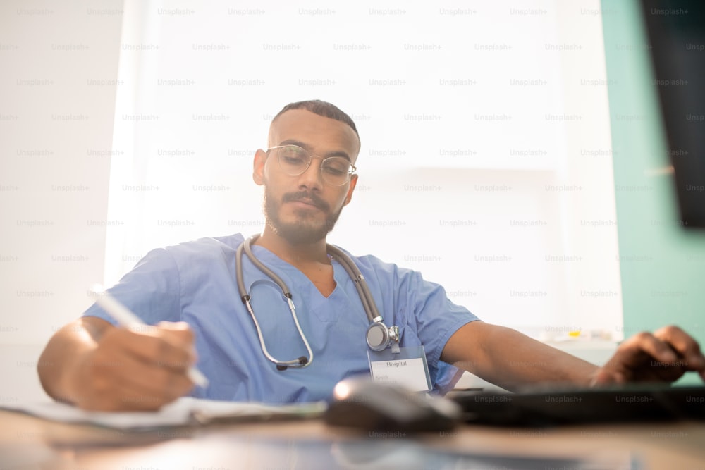Bearded serious doctor or intern in uniform making working notes while sitting by table in front of computer in clinics