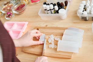 Hands of woman chopping white hard mass on wooden board while making handmade soap in studio